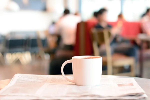 Coffee cup over newspaper on the table with people in coffee shop as blur background