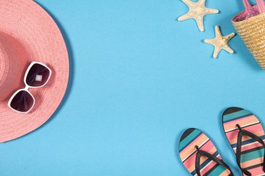 Mavi arkaplan, pembe şapka, güneş gözlüğü, parmak arası terlik ve yüksek açılı plaj aksesuarları.