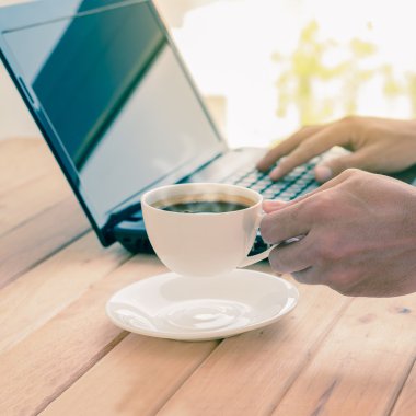 Kahve fincanı tutan ve bir dizüstü bilgisayar üzerinde ahşap masa yazarak adam. 