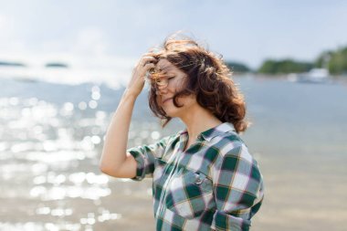 Sağlıklı bir kadının portresi. Yazın açık havada temiz hava soluyor. Arka planda deniz ve gökyüzü var. Kıvırcık saçlı genç kız güneşli bir bahar öğleden sonrasında parkta.