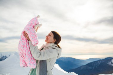 Anne kucağı yeni doğmuş bebeğe karlı yamaçta sarılıyor, mutlu anne kışın güneşli dağda küçük kız çocuğu kucaklıyor, sevgi ve ilgi gösteriyor, annelik izninin tadını çıkarıyor, çocuk bakımı kavramının tadını çıkarıyor. Kopyalama alanı ile