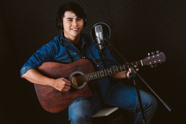İyi görünümlü Asyalı erkek şarkıcı akustik gitar çalarken stüdyoda gitar çalarken mikrofon ve pop kalkan kullanarak şarkı kaydediyor ve kulaklık takıyor..