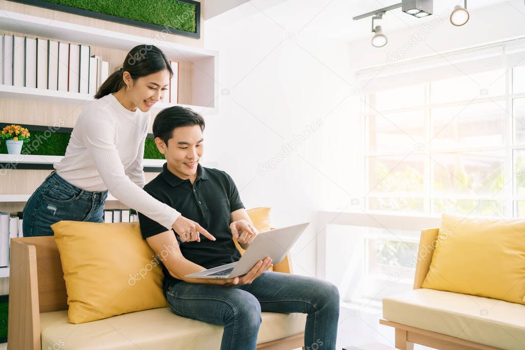 Asian male sitting and female standing back of him while working, pointing on the laptop together. Businessman working on laptop at home. Good opportunity comes with a surprise.