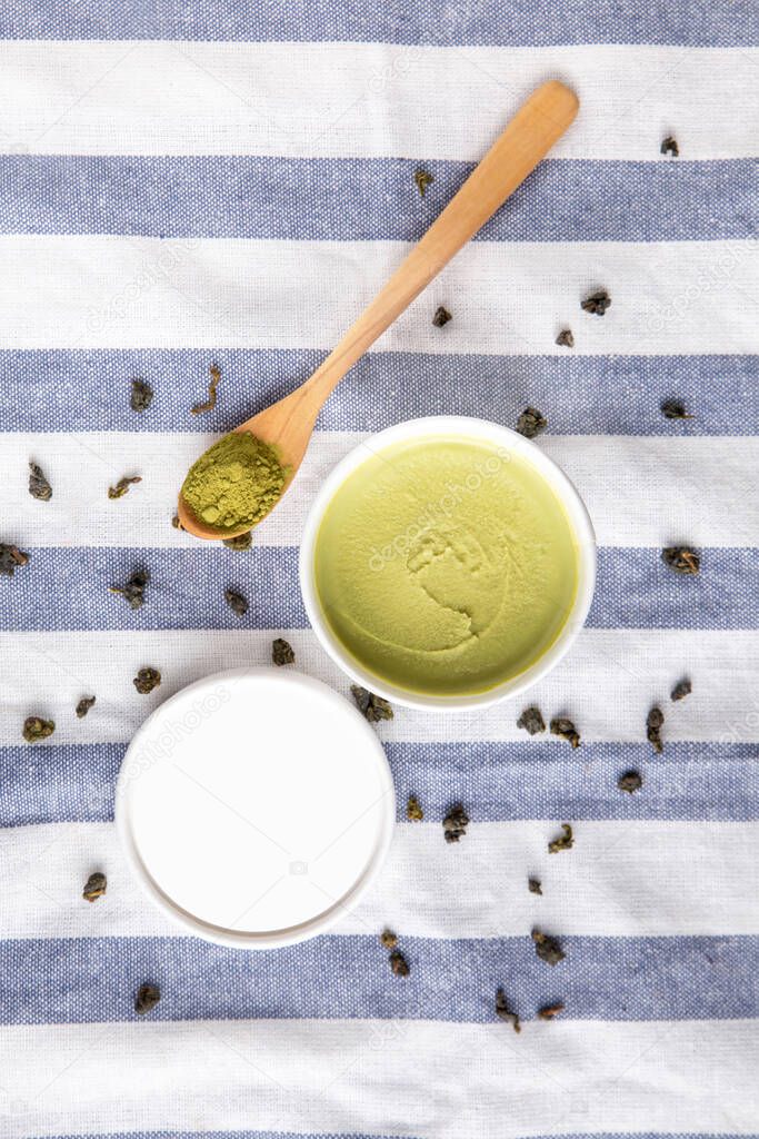 Top view of green tea matcha ice cream in a white paper cup with matcha powder on a wooden spoon on a blue and white stripe fabric with dry green tea leaves.