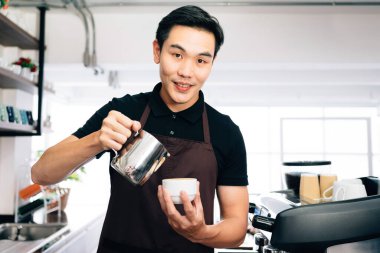 Genç erkek barista kameraya bakıyor, önlük giyiyor Latte Art yapmak için sıcak sütü sıcak espresso koyu kahvesine dolduruyor..