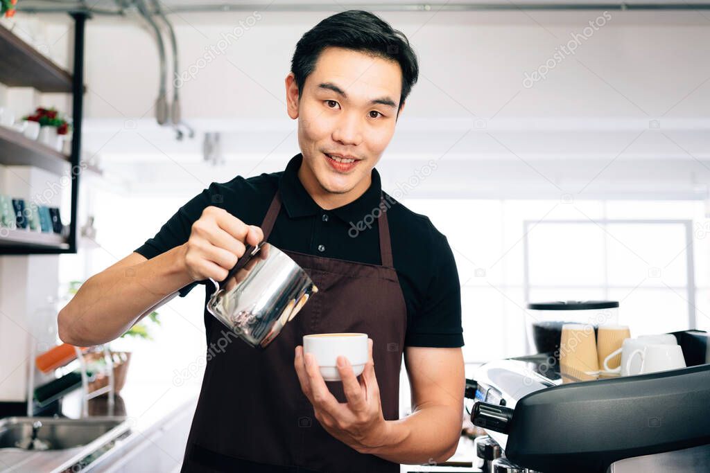 Young male barista looking at the camera, wearing an apron pouring hot milk into hot espresso black coffee for making Latte Art.