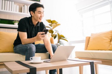 Asyalı bir erkek cep telefonu kullanırken ve masada dizüstü bilgisayarla çalışırken dışarıya bakıyor. Evde çalışan sıradan bir iş adamı, ofiste rahat bir yerde çalışıyor..