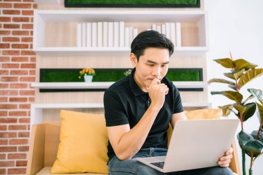 Sıradan giyinen Asyalı erkek, çalışırken ve kanepede otururken dizüstü bilgisayarla ilgilenir ve stres hisseder. İş adamı evde dizüstü bilgisayarla sorun yaşıyor. İş dünyasındaki sorun sürecinin çözümü.