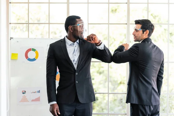 白人とアフリカのビジネスマンは顔の盾を身に着けていて コロナウイルスの流行の間にビジネスミーティングで握手する代わりに肘のバンプで挨拶する — ストック写真