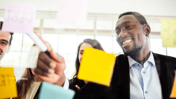 新しいアイデアを見つけるためにガラスの壁にカラフルな粘着紙のノートを使用して同僚と笑顔アフリカのビジネスマンブレインストーミング会議のクローズアップ アジャイル手法を用いてビジネスを行う — ストック写真