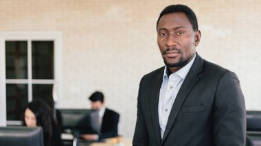 Kendine güvenen Afrikalı Amerikalı işadamı, arka planda masasında çalışan bulanık çalışanlarıyla ofiste duruyor. Afrikalı Amerikalı iş adamı portresi.