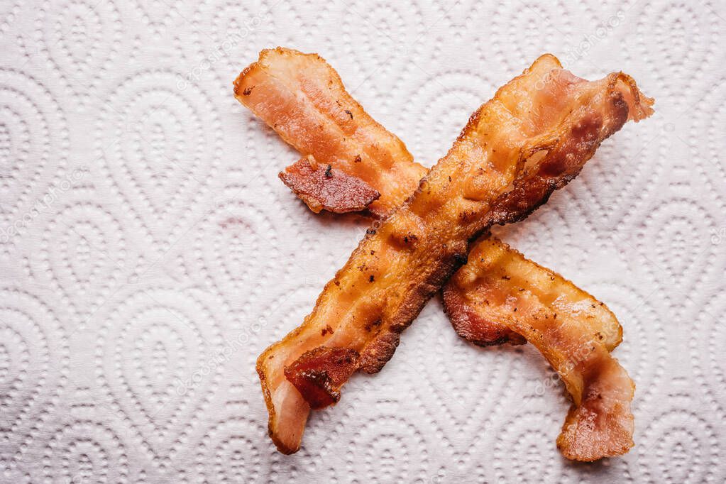 Top view of crispy bacon strips that put in the shape of a cross place on oil-absorbing tissue paper with copy space. Keto diet snack. Unhealthy greasy snack.