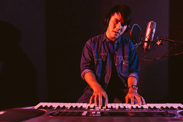 Male playing an electric keyboard while wearing headphones. Recording songs by using a studio microphone and pop shield on the microphone in blue and red light with copy space.
