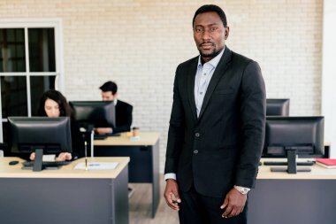 Kendine güvenen Afrikalı Amerikalı işadamı, arka planda masasında çalışan bulanık çalışanlarıyla ofiste duruyor. Afrikalı Amerikalı iş adamı portresi.