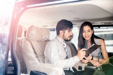 Araba galerisi. Asyalı satıcı (kadın) teslim etmeden önce plastik kaplı koltuklu yeni arabanın içindeki orta doğu müşterisiyle listeyi kontrol ediyor. Otomotiv Kiralama ve Pazarlama Şirketi.