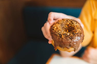 Sarı gömlekli bir kadın modern bir kafede fındıklı ev yapımı çikolatalı çörek tutuyor. Kadın yaşam tarzından zevk almak. Eline ve donutuna odaklan..