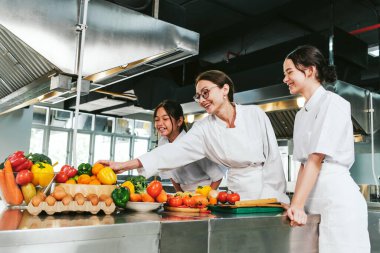Üniformalı şefler aşçılık dersleri sırasında paslanmaz tezgahtan taze sebze seçerek işbirliği yaparlar. Gülümseyen personel profesyonel bir okul mutfağında takım çalışması, eğitim ve işyeri eğitimi gösterir.