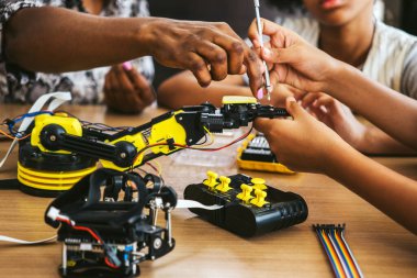Bir STEM atölyesi sırasında araç ve telleri kullanarak sarı bir robotik kol kitini bir çalışma tezgahında birleştirmek için işbirliği yapan eller, takım çalışması, mühendislik ve pratik robotik eğitimi gösteriyor.