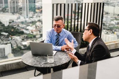 Takım elbiseli iki işadamı, şehrin siluetiyle çatıda buluşup dizüstü bilgisayarın planlarını tartışıyor. Açık hava girişimleri ortaklık, işbirliği, liderlik, takım çalışması, beyin fırtınası ve anlaşma gösteriyor.