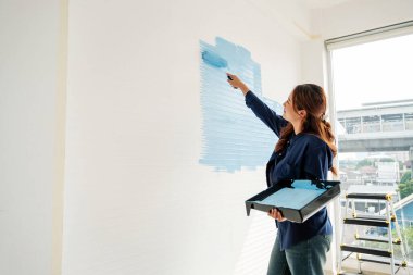Kadın, beyaz iç duvarı parlak pencerenin yanındaki tepsiyi tekerlekli sandalyeyle açık maviye boyuyor. DIY daire yenileme sahnesi boyama, yenileme, yenileme, fırçalama ve ev geliştirme projelerini gösteriyor..