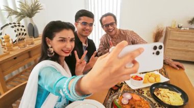 Şenlikli giyinmiş üç Hintli arkadaş, rahat bir evde yemek dolu bir yemek masasında neşeli bir selfie çekiyorlar, keyifli bir toplanma ve paylaşılan bir yemek eşliğinde kutlama yapıyorlar..