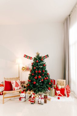 Parlak oturma odası dekore edilmiş Noel ağacı, kırmızı ve beyaz süsler, hediyeler, ve pencerenin yanında şenlikli bir dekor, rahat bir tatil ortamı yaratıyor..