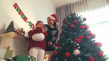 Noel Baba şapkalı anne ve kız Noel ağacının yanında hediyeler ve konfeti ile sıcacık bir oturma odasında gülüyorlar, tatili parlak doğal ışık altında kutluyorlar. Gerçek zamanlı 4k video