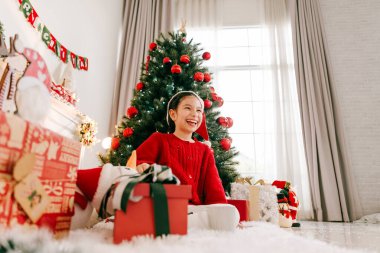 Noel Baba şapkalı gülümseyen kız Noel ağacı süsleri, hediyeler ve hediyelerle süslü bir halının üzerinde oturuyor. Konforlu oturma odası sahnesi, kış tatili kutlamalarını ve şenlik ruhunu yakalıyor..