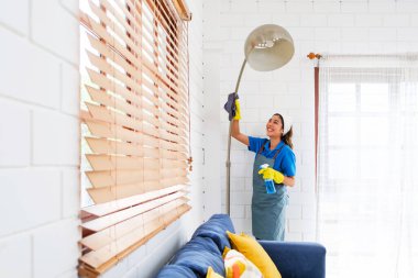 Gülümseyen ev temizleyicisi parlak bir oturma odasında uzun bir lambanın tozunu alır, koltuk ve pencere perdelerinin yanında eldiven ve önlük giyer..