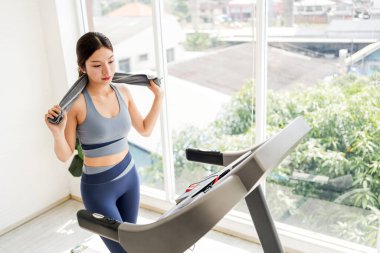 Genç bir kadın, parlak spor salonunda koşu bandı çalışmasından sonra havluyla serinliyor. Sahne kardiyo eğitimi, koşu, koşu, spor, egzersiz, sağlık ve yeni yıl çözünürlük motivasyonunu yansıtıyor.