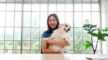 Evde elinde sevimli bir Corgi köpeği olan neşeli genç bir kadın gülümseyerek kameraya bakıyor, büyük bir pencerenin önünde oturuyor, neşeyi, kaynaşmayı ve evcil hayvan dostu bir yaşam tarzını temsil ediyor..