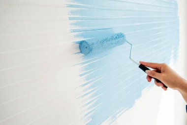 El, bir ev yenilemesi sırasında beyaz desenli duvara açık mavi boya sürmek için boya silindiri kullanıyor.