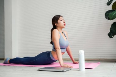Asyalı genç bir kadın evdeki pembe yoga minderinde poz veriyor, bir dizüstü bilgisayar ve su şişesinin yanında derin derin nefes alıyor parlak, minimalist bir odada bir bitkiyle birlikte. Yeni yıl kararı, kendinize kıyak yapın..