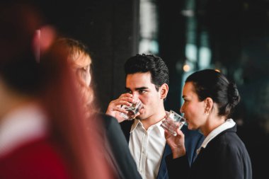 Resmi giyinme ağı içindeki profesyoneller kapalı bir etkinlikte su içip loş bir salonda sohbet ediyorlar..