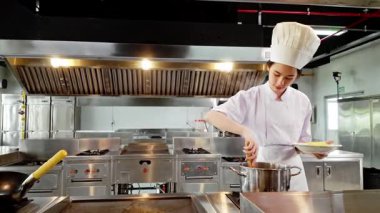 Beyaz aşçı üniformalı genç bir öğrenci profesyonel bir mutfaktaki kızgın bir tencerede sıcak makarna kaplıyor, aşçılık eğitimi sırasında pratik pişirme becerileri sergiliyor..