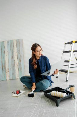 Bir ev boyama projesi sırasında yerde oturan kişi, fırça tutarken dizüstü bilgisayarı kontrol ediyor; boya rulo tepsisi, mezura ve yakınlardaki parlak bir iç mekanda merdiveni kontrol ediyor