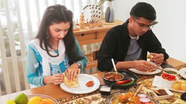 Üç Hintli yemek masasında ev yapımı bir yemeği paylaşıyor. Parlak, rahat bir odada tabağı uzatıyor. Beraberliği, konukseverliği ve günlük ev yemeklerini vurguluyor..