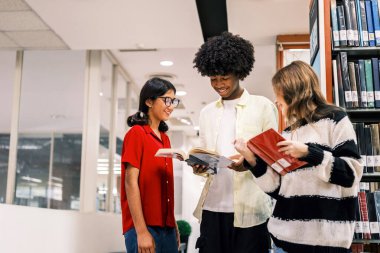 Üç neşeli öğrenci parlak bir kütüphane koridorunda duruyorlar, uzun rafların yanında gülümsüyor ve kitapları karşılaştırıyorlar, gençlik eğitimini, öğrenme kaynaklarını ve akademik bağlılığı simgeliyorlar..