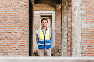 Güvenlik yeleği giymiş kendine güvenen bir adam kısmen inşa edilmiş tuğla bir yapıda duruyor, kameraya bakıyor, inşaat sürecini, teftişi ve inşaat sektörünü sembolize ediyor..