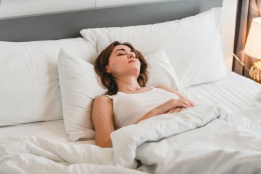 Genç bir kadın, beyaz çarşaflı, parlak, rahat bir yatak odasında huzurlu bir şekilde dinleniyor. Sakin, destekleyici bir ev ortamında huzurlu bir uyku, rahatlık ve sağlık anı yakalıyor..