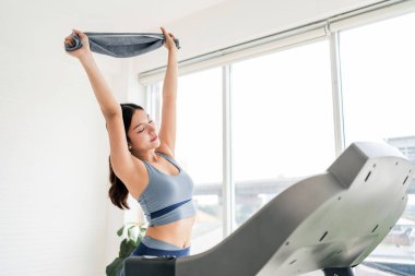 Antrenman kıyafetleri içinde bir kadın koşu bandının önünde havluyla esniyor, büyük pencerelerden gelen doğal ışıkla banyo yapıyor, sağlık ve pozitifliği yeni bir spor salonunda kucaklıyor..