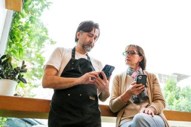 Kafe sahibi ve işletme danışmanı akıllı telefonlar kullanarak pazarlama veya finansmanı tartışıyor. Her ikisi de gülümsüyor ve meşgul, takım çalışmasını, stratejiyi ve modern küçük iş ortaklığını sembolize ediyor..