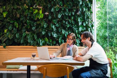 İki meslektaş, kafe sahibi ve finans danışmanı, bir kafede dizüstü bilgisayarla kapalı yeşil bir duvarın yanında iş tartışırken, modern, gündelik bir çalışma alanında danışmanlık veya küçük işletme planlama önerisinde bulunuyorlar..