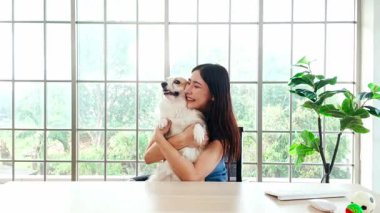 Evde elinde sevimli bir Corgi köpeği olan neşeli genç bir kadın gülümseyerek kameraya bakıyor, büyük bir pencerenin önünde oturuyor, neşeyi, kaynaşmayı ve evcil hayvan dostu bir yaşam tarzını temsil ediyor..