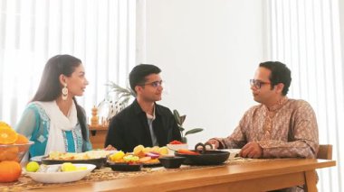 Üç Hintli yemek masasında ev yapımı bir yemeği paylaşıyor. Parlak, rahat bir odada tabağı uzatıyor. Beraberliği, konukseverliği ve günlük ev yemeklerini vurguluyor..