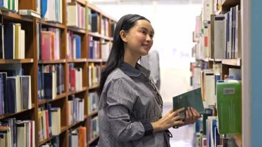 Genç Asyalı kadın, parlak bir kütüphane koridorunda kitapları incelerken gülümsüyor, uzun raflar arasında yeşil bir kitap tutuyor, okuma, öğrenme, edebiyat ve akademik keşif temalarını yakalıyor.