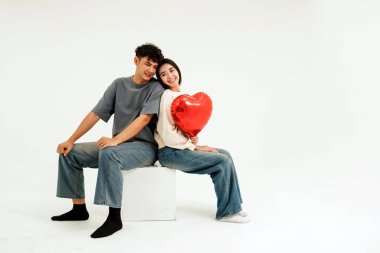 Genç bir çift sırt sırta oturup gülümserken kadın elinde kırmızı kalp şeklinde bir balon tutuyor. Temiz beyaz bir arkaplan, sevgililer günü için sevgi, birlik, mutluluk ve ilişki içeriği temalarını yakalar..