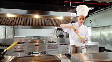 Beyaz aşçı üniformalı genç bir öğrenci profesyonel bir mutfaktaki kızgın bir tencerede sıcak makarna kaplıyor, aşçılık eğitimi sırasında pratik pişirme becerileri sergiliyor..