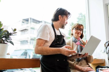 Bir barista ve bir müşteri büyük pencereli ve kapalı mekanlı bir kafede dizüstü bilgisayarda bir şey tartışıyorlar..