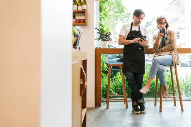 Barista ve müşteri parlak, modern bir kafede maske takıyor, bir pencere barına sipariş vermek ya da ödemek için akıllı bir telefon görüyor, rahat iç mekan ayrıntıları ve şehir manzaralı.